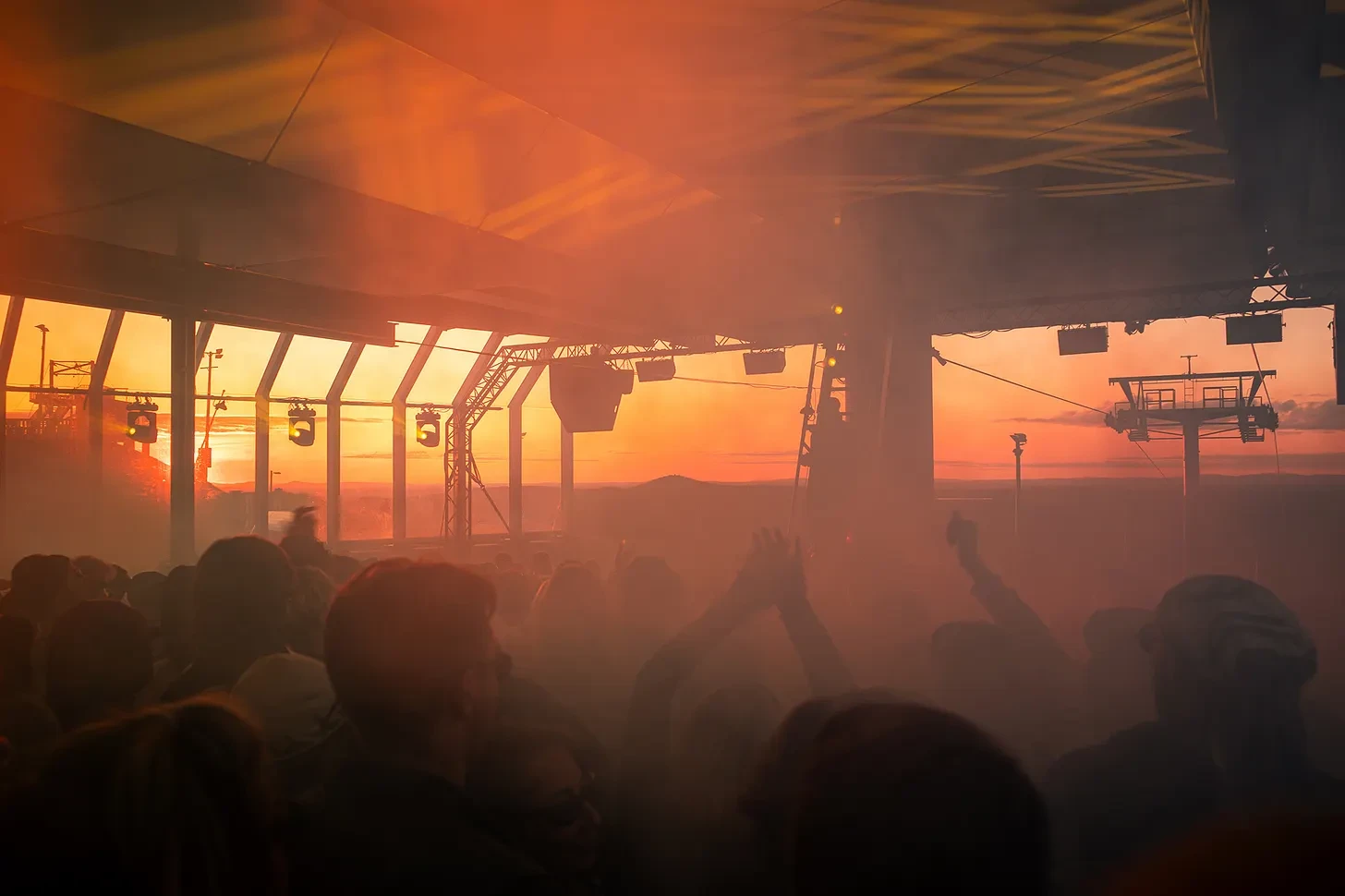 The crowd at Solstice in haze and sunset light under the roof, with the fells and a ski lift beyond
